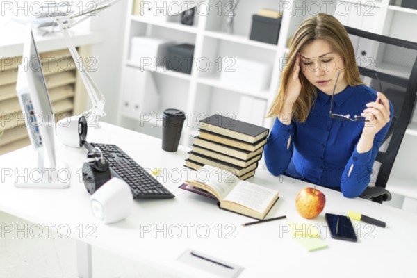 A young girl is sitting at a computer desk, holding glasses in her hand and rubbing her eyes. Before the girl lies an open book and an apple
