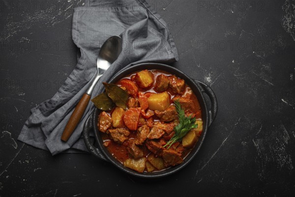 Beef meat stew with potatoes, carrot and delicious gravy in black casserole pot with bay leaves with spoon on black dark rustic concrete background from above, food photography, food photography