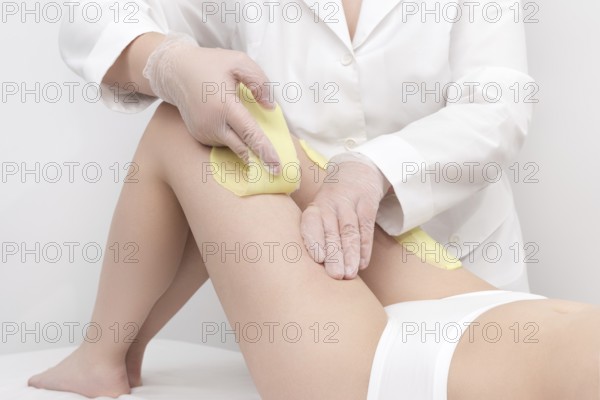 Unrecognizable beautician in white coat removal hair on women legs. Depilation procedure with green hot wax in professional beauty salon while woman lying down on couch. Part of photo series