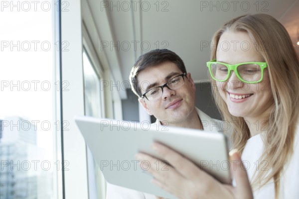 Man and woman in glasses are standing by window and watching something in tablet PC