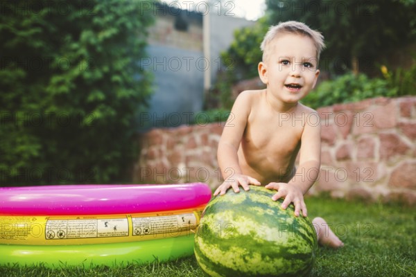 Toddler cute Caucasian white European race boy sitting hugging holding huge watermelon and smiling on background of green graß, backyard of the house and a round children pool on a sunny summer day