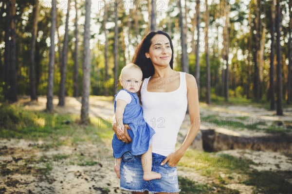 Young beautiful fashionable mother with long brunette hair holding a daughter's blonde with blue eyes one year of birth in a coniferous forest in a summer park. Wearing jeans clothes
