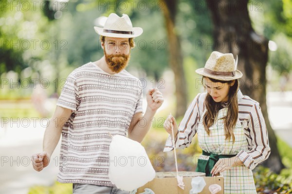 Theme is a family small business cooking sweets. A pair of lovers A young man and woman trader in the hat the owner of the outlet makes candy floß, fairy floß or Cotton candy in the park in summer