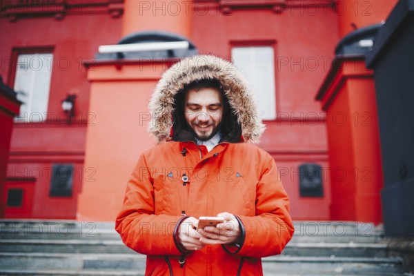Handsome young male student with toothy smile and beard stands on red wall background, facade of educational institution in red winter jacket with hood with fur, Uses finger on screen of mobile phone