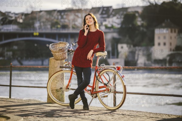 A young female traveler enjoying stands next to a bicycle near a river in the city of Basel. Traveling in Switzerland. Young caucasian woman talking by cell phone on bridge in european city