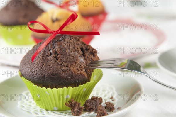 Lovingly decorated muffins on a coffee table. In the foreground, a chocolate muffin with a red bow and green sleeve that has already been tasted. A cake fork is leant against the side. Crumbs lie on the plate. A cake platter with more muffins in the background. Bright light and friendly atmosphere. In the foreground a chocolate muffin already tasted with red ribbon and a green collar. A cake fork is leaning sideways. Crumbs on his plate. In the background a cake plate with more muffins