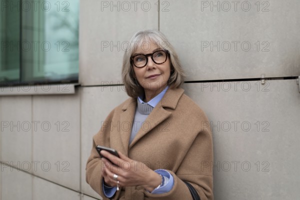 Eine reife Frau in einem hellbraunen Mantel steht an einem modernen Gebäude und überprüft ihr Telefon, während sie einen Moment der städtischen Ruhe genießt