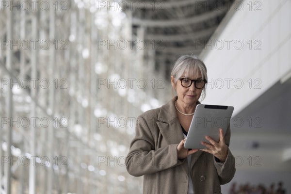 An elderly businesswoman stands confidently in a contemporary office space, engaging with a tablet. She showcases her expertise and professionalism amid a bustling work environment