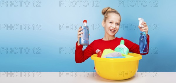 Isolated on blue, cheerful girl in red pullover with fair-haired updo, holding in both hands a spray and a cleaner from a lemon washbowl with multicolored detergents and rags. copyspace