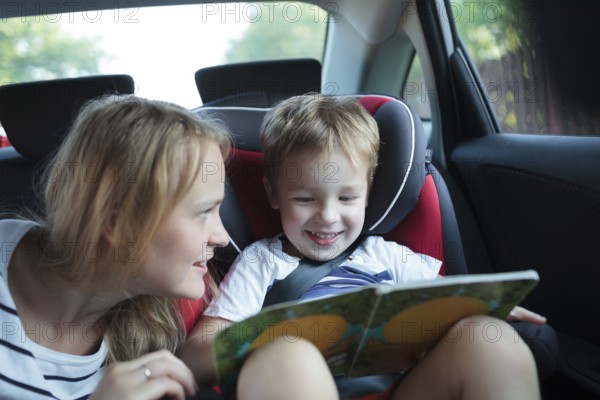 Little boy in a car sitting in child safety seat looking at the pictures in the book, mother watching him