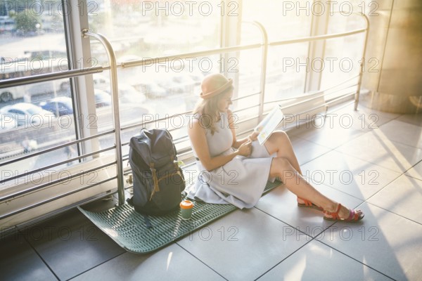 Waiting, delayed transport in the terminal of the airport or train station. Young caucasian woman in dress and hat sits on tourist rug with backpack near window to room and looks into tourist map