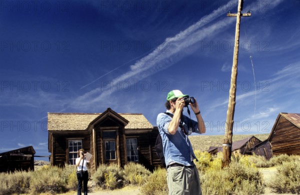 Two tourists photographing and marvelling in front of a wooden house in Bodie. Colour photo, landscape format. Two photographing and marvelling tourists in front of one of the old wooden houses of Bodie. Colour photo, horizontal format