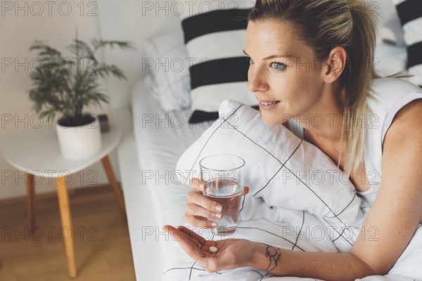 Smiling young caucasian woman taking her morning supplements, vitamin pills with a glass of water at home.