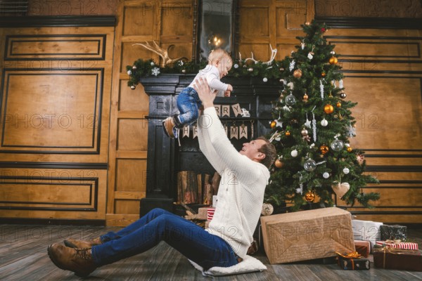 Happy father and son at home near Christmas tree. New Year and Christmas theme. Holiday mood. Family winter holidays concept. Father raises son in arms sitting on floor near Christmas tree and gift