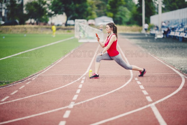 Sports fitness Caucasian women doing warm-up before exercise. Athlete stretching legs doing warm-up before training. Jogging in the city at the stadium. Girl uses headphones handset phone sportswear