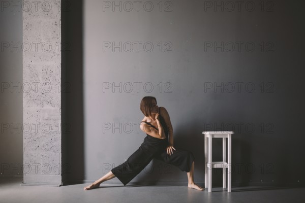 Social theme of female loneliness is pain suffering. Abastration of male domestic violence. Young beautiful Caucasian woman in black clothes, strange pose, white chair big dark room. Fear and despair