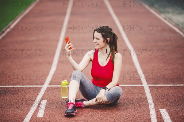 Young woman athlete at the stadium sporting lifestyle, sitting on the track, taking selfie photo on a smartphone, smiling joyful in her ears earpiece, near a bottle flask with water
