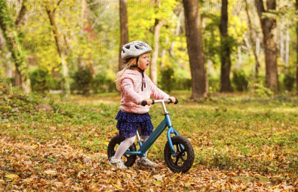 Side view preschool girl in protective helmet riding blue balance bike at the autumn park. Lovely girl in skirt on bike among fallen leaves