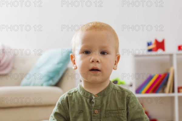 Portrait of a baby boy who is enchanted looks at the camera. The blond boy is sitting in the middle of the playroom with his mouth open
