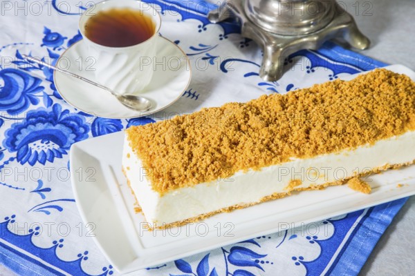 Professional bakery. Exquisite white cheesecake, sprinkled with sweet orange crumbs. The background is samovar and porcelain white cup with hot tea on blue kitchen towel
