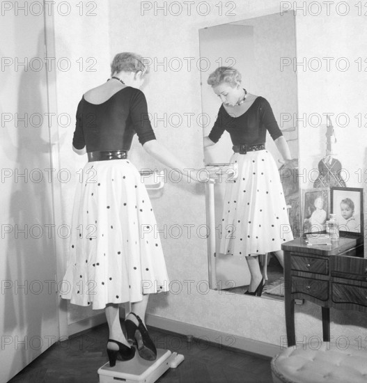 1950s fashion. A young woman is standing on one leg on the scale trying to be as light as possible. Pictures actress AnnaLisa Ericson, 1913-1911. Sweden 1954. Photo Kristoffersson ref BM74-10