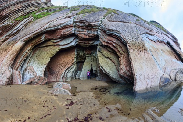 Picturesque huge cave on the beach. Low tide in the Atlantic. Flysch - unique landscape of underwater rocks exposed at low tide. Woman in pink jacket photographs Playa Zumaya