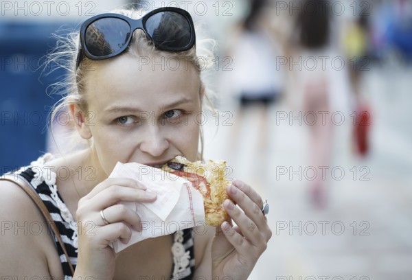 Young woman with sunglasses on her head eating fast food in the street and looking around being thoughtful