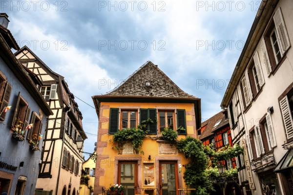 Characteristic half-timbered house with colourful shutters and floral decorations under a cloudy sky. Alsace France