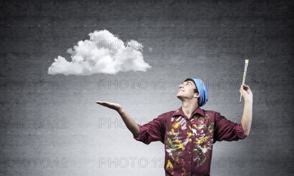 Young artist drawing realistic white cloud. Male painter in dirty shirt and bandana with paintbrush on grey wall background. Creative hobby and artistic occupation. Art classes concept with copy space