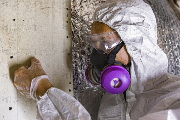 A man wearing protective breathing equipment is viewed closeup, inspecting signs of mold and mildew inside the walls of a family home