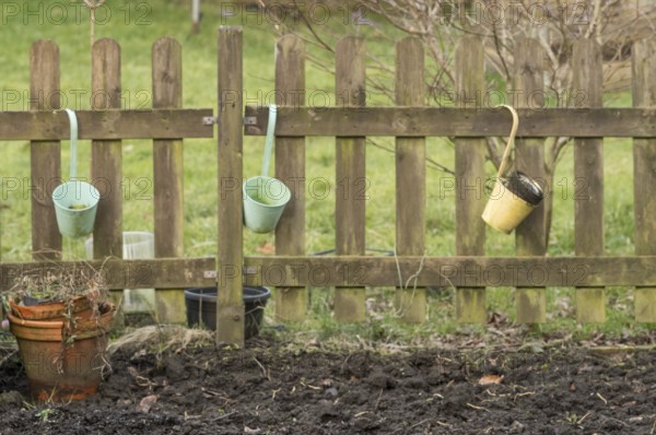 Three empty buckets hanging on a weathered wooden fence in a garden, with a terracotta pot and bare soil in the foreground