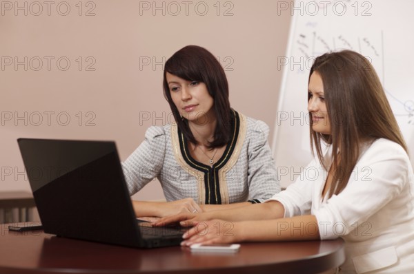 Two attractive stylish young women sitting at a table with a laptop computer reading something on the screen together while browsing the internet