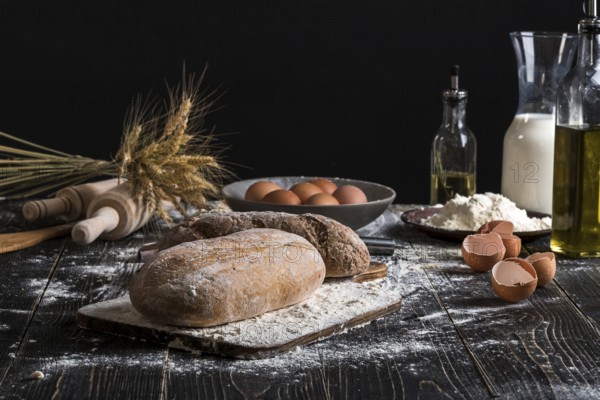 Beautiful still life with different kinds of bread, grain, flour on weight, ears of wheat, pitcher of milk and eggs on black background