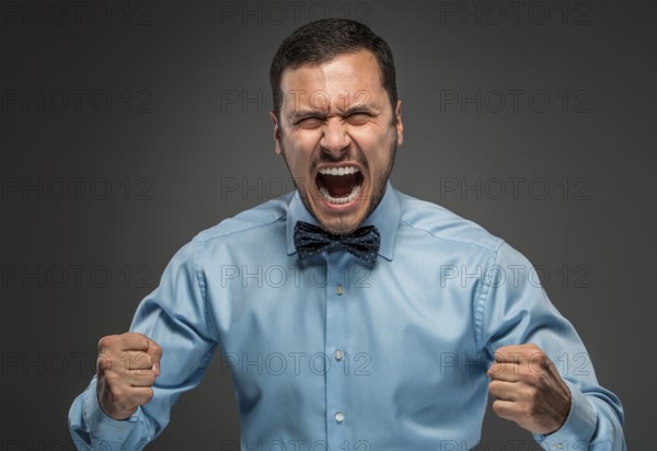 Portrait of angry upset young man in blue shirt and butterfly tie with fists up yelling isolated on gray studio background. Negative human emotion, facial expression. Closeup