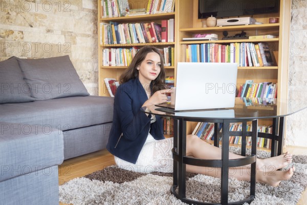 Beautiful young woman, dressed elegantly, sits on the carpet in the coziness of her home, enjoying a cup of coffee, while using a laptop
