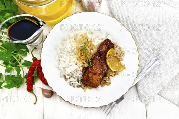 Pink salmon with honey, lemon juice and soy sauce, boiled rice, slice of lemon and sprig of thyme in a plate, hot pepper, garlic, parsley, napkin and fork on wooden board background