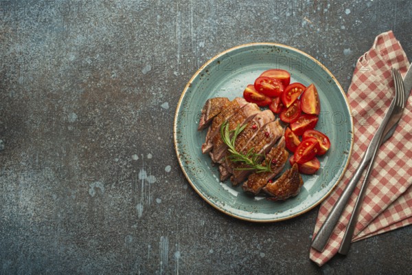 Delicious roasted sliced duck breast fillet with golden crispy skin, with pepper and rosemary, top view ceramic blue plate served with cherry tomatoes salad, concrete rustic background, copy space, food photography, food photography