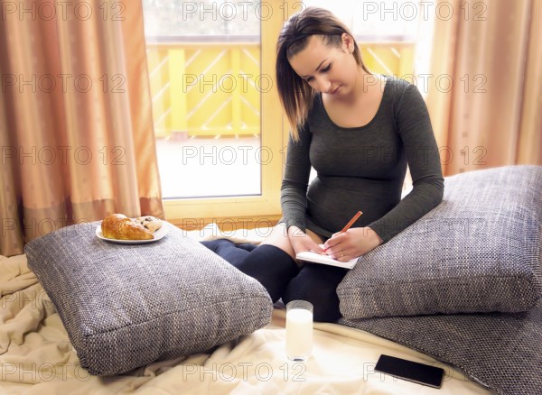Beautiful pregnant woman sits comfortable on pillows, in front of big windows, with snacks nearby and writes in a notebook