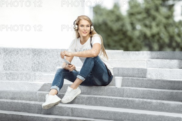 Happy young woman with vintage headphones and a cup of coffee to go, surfing the internet on a tablet PC, listening to music and sitting on a staircase in front of an urban background