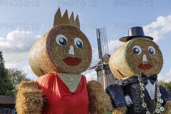 Two tall straw men with hats and crowns stand in front of a windmill under a blue sky with clouds, weseke, münsterland, germany