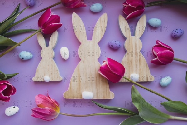 Easter eggs and wooden bunnies with pink tulips on violet pastel background. Flat lay composition, top view. Decoration minimalist modern design template. Floral layout Concept of creative spring holiday mock up