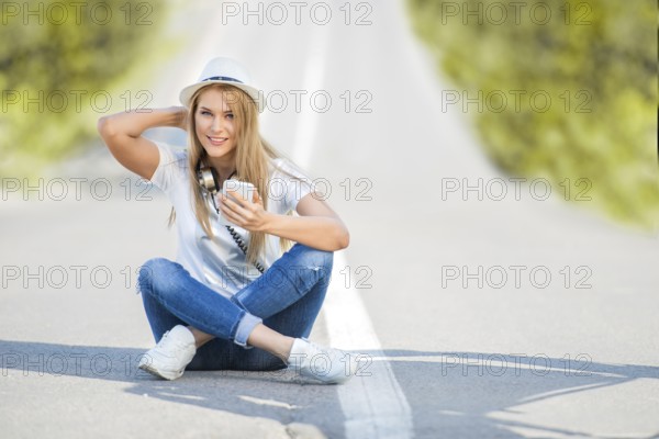 Happy young woman with vintage headphones around her neck, surfing the internet on her smartphone and sitting on a divider against the backdrop of a street