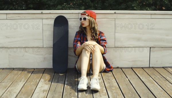 Stylish young woman sitting with skateboard in casual clothes looking away, modern teenage girl posing in the city