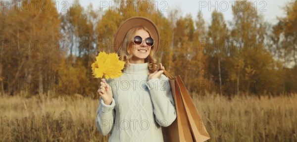 Autumn portrait of stylish happy young woman with shopping bag and maple yellow leaves, fashionable girl smiling wearing glasses, hat, sweater outdoors in park, warm fall season