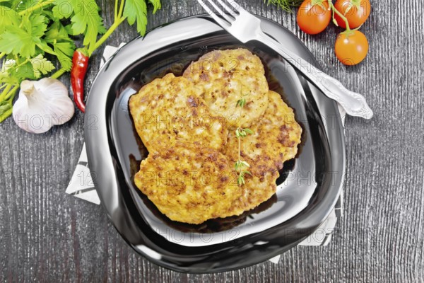 Fritters of minced meat in a plate with a fork on napkin, garlic, parsley and hot pepper on black wooden board background from above