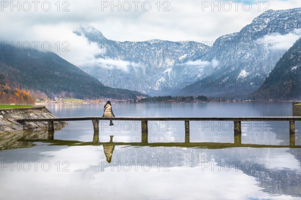 Woman sitting on a wooden bridge over the lake Hallstatter, admiring the peaks of the Austrian Alps and their reflection in the water, in Hallstatt, Austria
