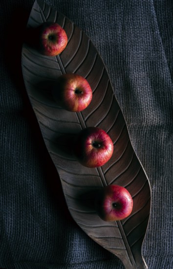 Red apples on a wooden tray in shape of a leaf, on a jute sack, in low light, above view