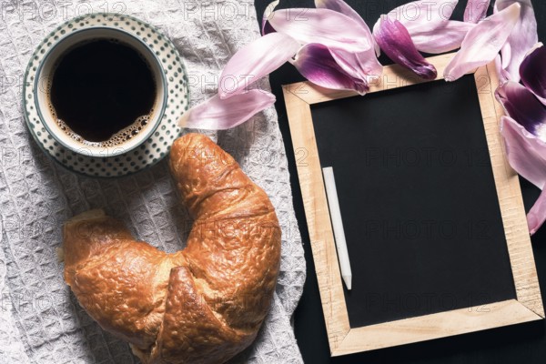 Cup of hot coffee and a fresh croissant on a retro kitchen towel, near a blank chalkboard, surrounded by pink tulip petals, on a black table