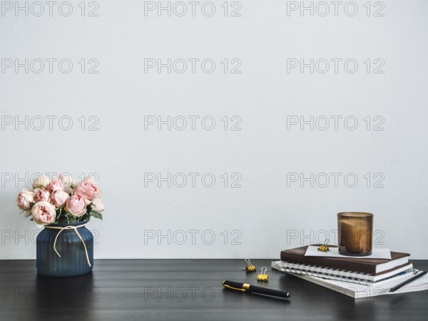 Home office desk with copy space. Pink flowers, scented candle, stack of books on black tabetop. Copy space for text. Home office desk workspace. Empty white room with wooden table