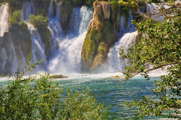 An impressive waterfall flows into a clear, green river, surrounded by lush vegetation and bright sunshine, Krka National Park Lozovac Dalmatia Croatia